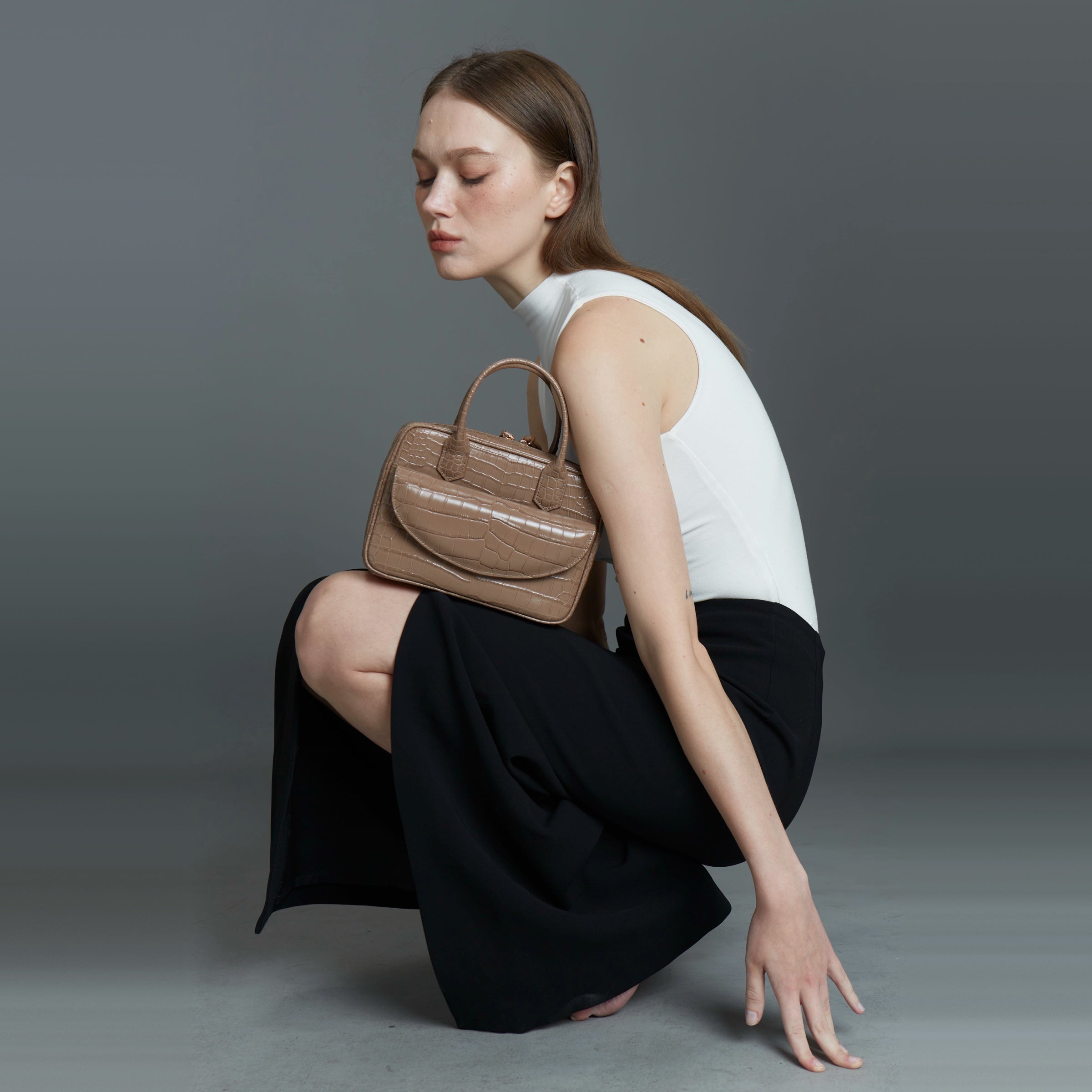 Model crouching in a white top and black skirt with a taupe croc-texture Klove handbag